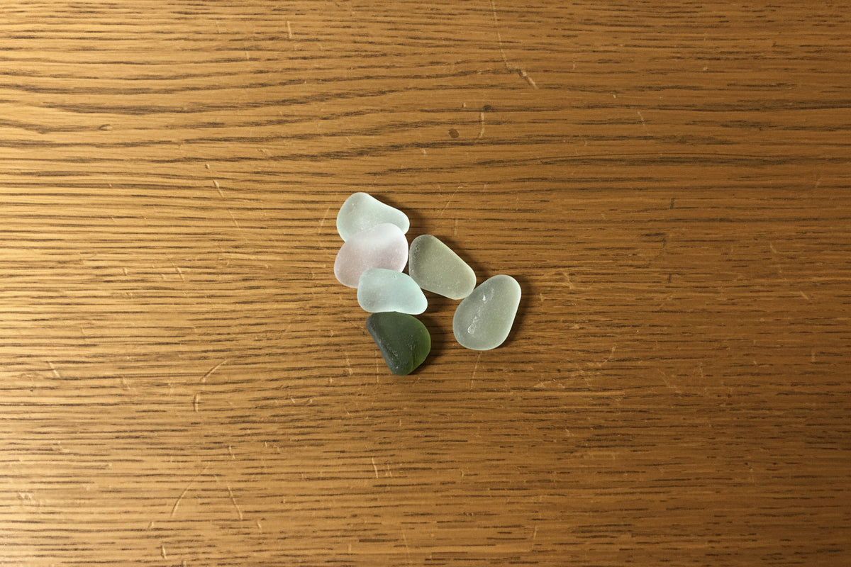 Five smooth, polished sea glass pebbles in pastel and muted colors, including pale green, frosted white, and dark olive green, arranged on a textured purple background.