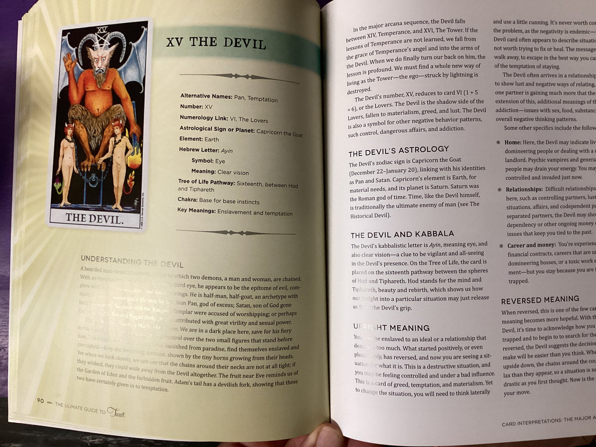 An open book showing a detailed page about 'XV The Devil' from a tarot card guide, held by a hand at the bottom. The page features a colorful illustration of the devil, astrological details, and text columns.